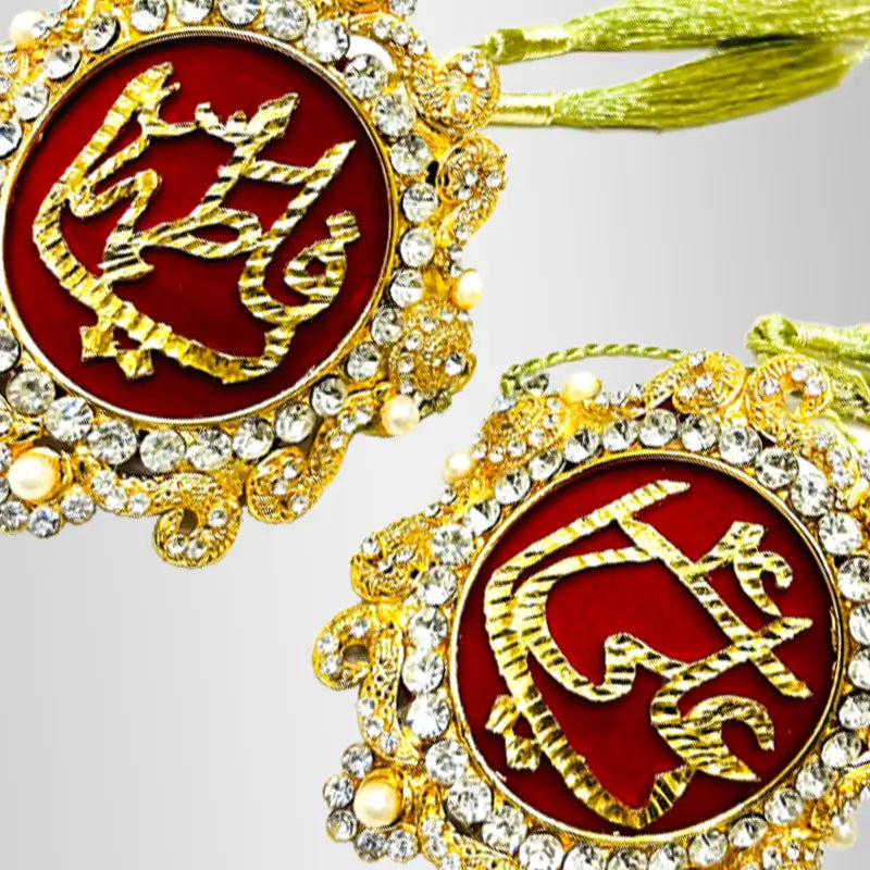 Two ornate gold-toned decorative ornaments with red circular inserts featuring gold Arabic calligraphy, surrounded by clear rhinestones and small pearl-like accents, with golden tassels visible at the top on a red velvet background.